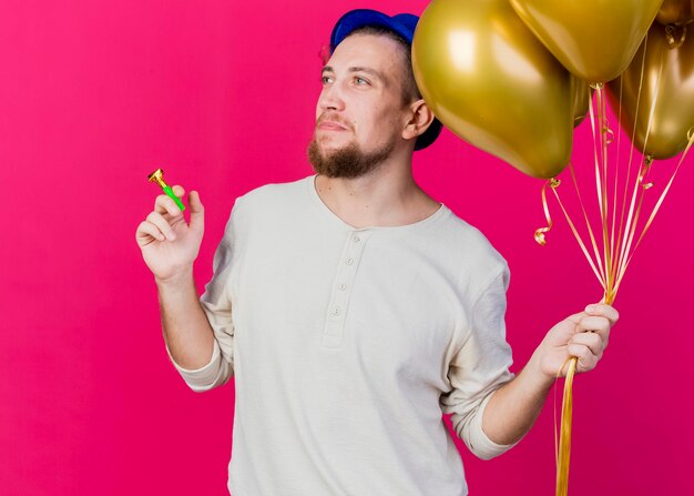 Young handsome slavic party guy wearing party hat holding balloons and party blower looking at side isolated on pink wall