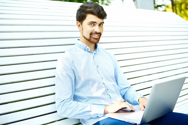 Young handsome smiling businessman model sitting on the park bench using laptop in casual hipster cloth