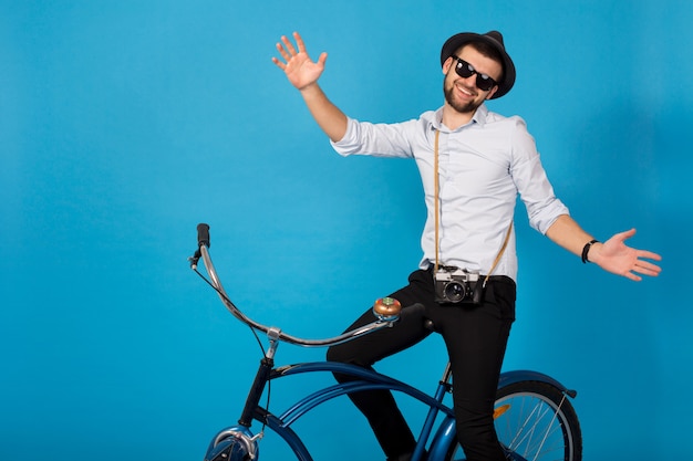 Free photo young handsome smiling happy man traveling on hipster bicycle, posing on blue studio background, wearing shirt, hat and sunglasses