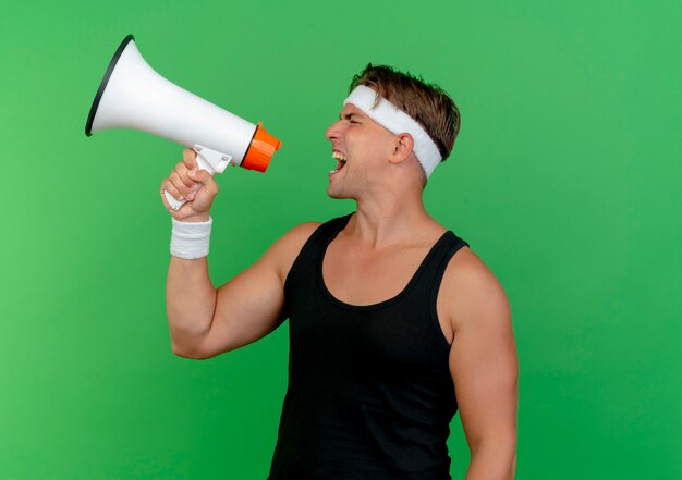 Young handsome sporty man wearing headband and wristbands shouting in loud speaker isolated on green background with copy space