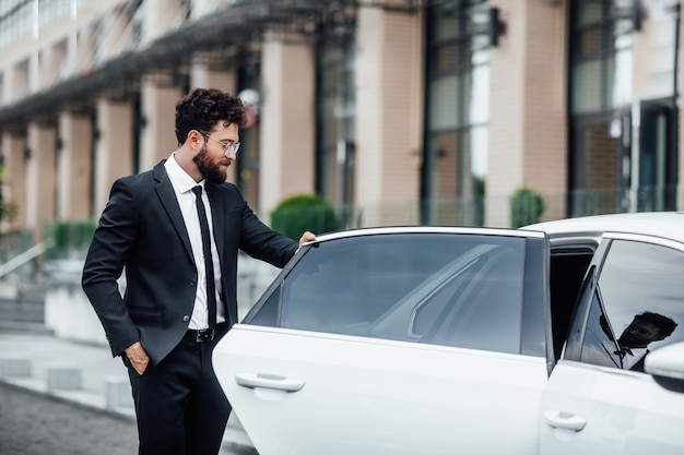 Free photo young handsome successful manager in black suit entering the rear seat of his car near a modern business center, on the street of big city