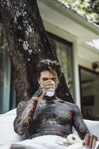 Free photo young handsome tattooed man having breakfast in bed in the open air outdoors in the garden