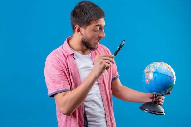 Free Photo young handsome traveler guy holding globe and looking at it using magnifying glass looking surprised standing over blue background