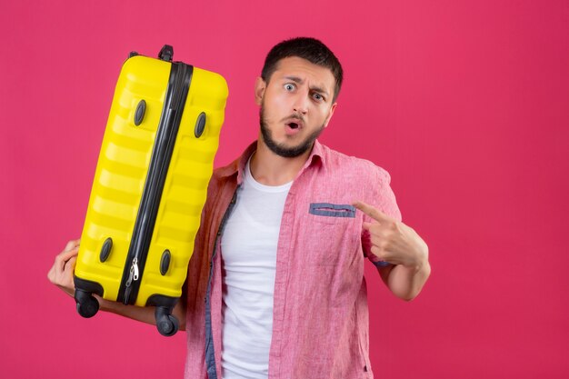 Young handsome traveler guy holding suitcase looking amazed and surprised pointing to himself standing over pink background