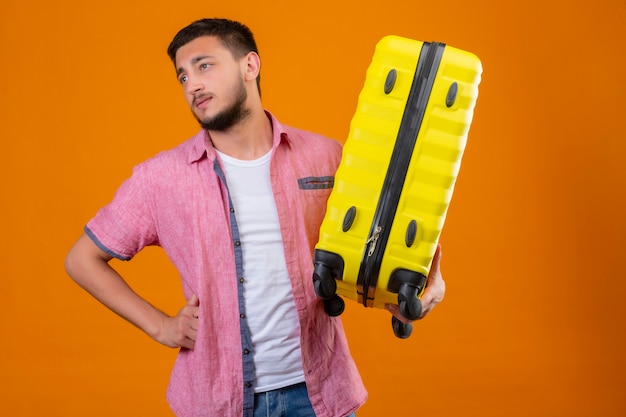 Free Photo young handsome traveler guy holding suitcase looking aside bored and tired with sad expression on face standing over orange background