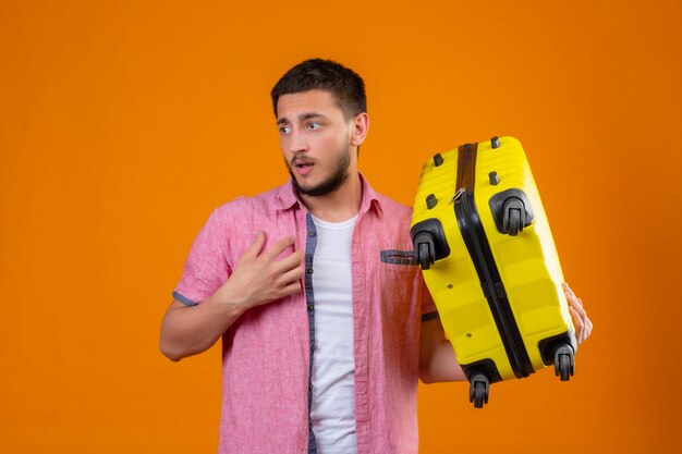 Young handsome traveler guy holding suitcase looking aside with sad expression on face unhappy standing over orange background