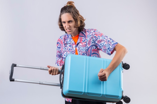 Young handsome traveler guy holding suitcase using as a guitar looking with angry expression standing over white background