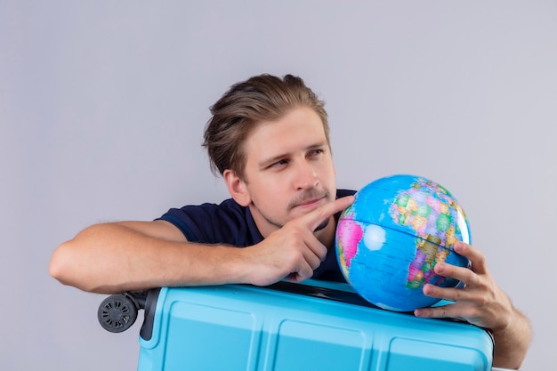 Young handsome traveler guy standing with suitcase holding globe looking aside with pensive expression thinking over white background