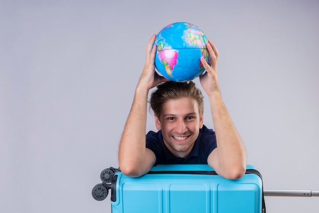 Young handsome traveler guy standing with suitcase holding globe looking at camera smiling cheerfully with happy face over white background