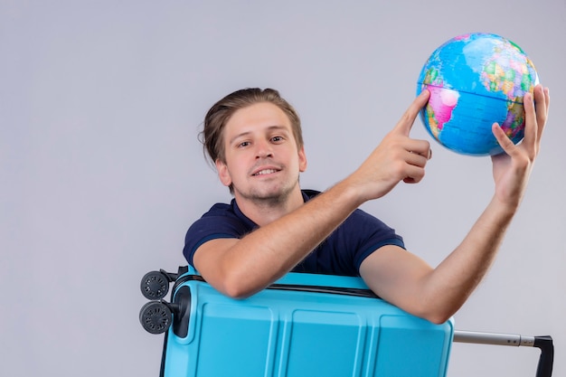 Young handsome traveler guy standing with suitcase holding globe looking at camera with confident expression smiling over white background