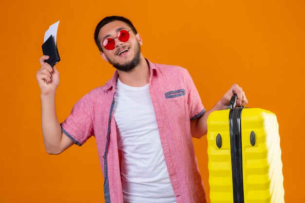 Young handsome traveler guy wearing sunglasses holding suitcase and air tickets looking confident self-satisfied smiling cheerfully ready to travel standing over orange background