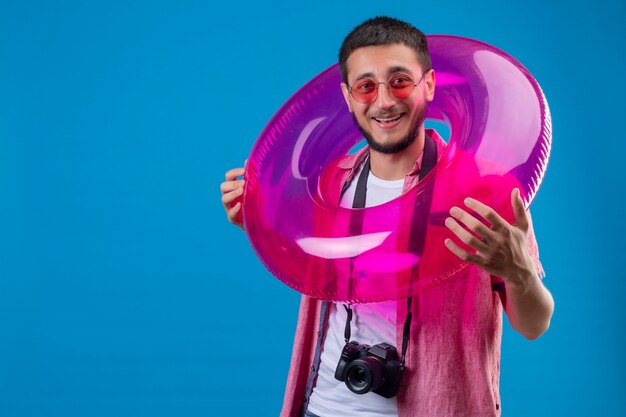 Young handsome traveler guy wearing sunglasses standing with inflatable ring looking at camera with happy face smiling cheerfully standing over blue background