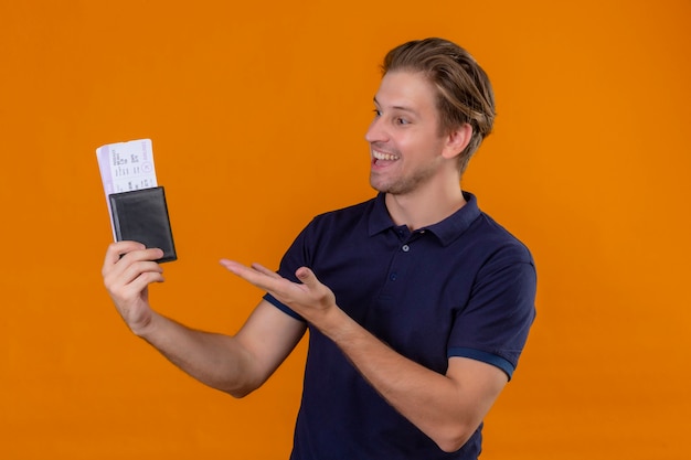 Young handsome traveler man holding air tickets presenting with arm of hand looking to them with smile on face over orange background