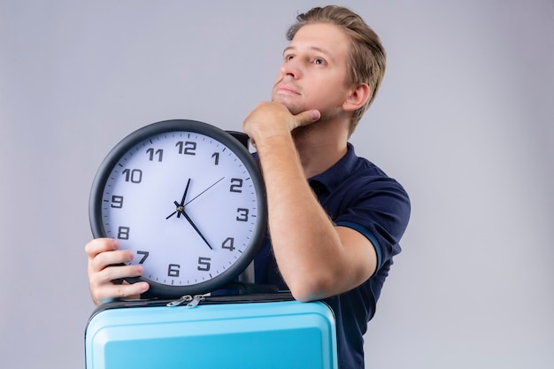 Young handsome traveler man holding clock with hand on chin looking away with pensive expression thinking over white background