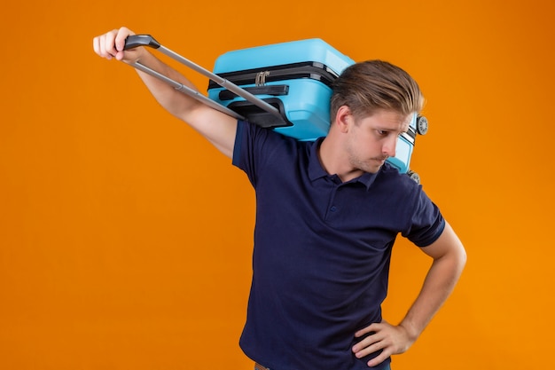 Young handsome traveler man standing with suitcase looking aside tired and bored over orange background