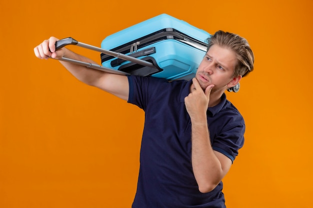 Free Photo young handsome traveler man standing with suitcase looking aside with hand on chin thinking having doubts over orange background