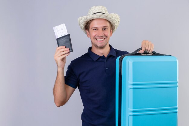 Young handsome traveler man in summer hat standing with suitcase holding air tickets looking at camera with happy face smiling cheerfully over white background