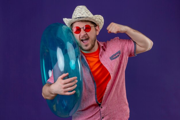 Young handsome traveler man in summer hat with inflatable ring raising fist