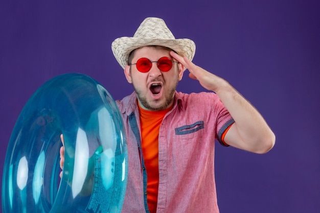 Young handsome traveler man in summer hat with inflatable ring with hand on head