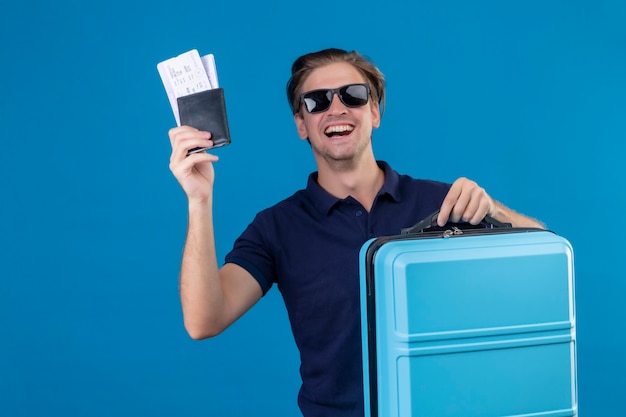 Young handsome traveler man wearing black sunglasses standing with suitcase holding air tickets looking at camera with happy face smiling cheerfully over blue background