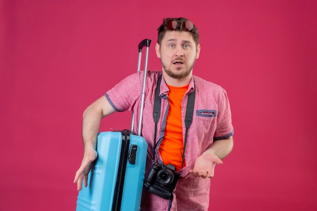 Free photo young handsome traveler man with camera standing with suitcase looking confused shrugging shoulders looking uncertain and confused