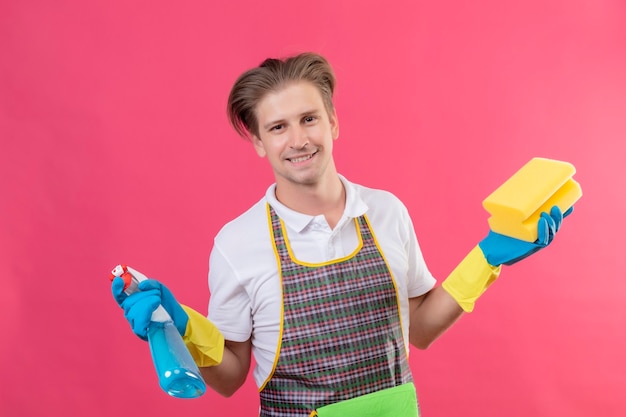 Young hansdome man wearing apron and rubber gloves holding cleaning spray and sponge with big smile on face happy and positive standing over pink wall