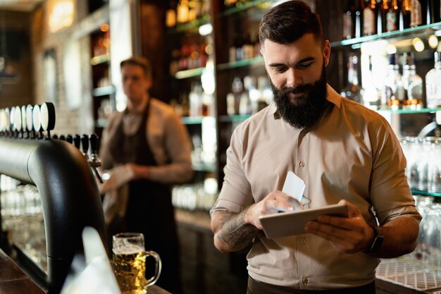 Young happy barista working in a bar and surfing the net on touchpad.