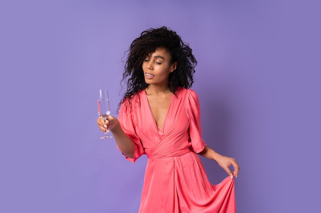 Young  happy  brazillian  female with  curly  hair in  pink stylish dress posing with glass  of 
champagne  over purple wall.  Party mood.