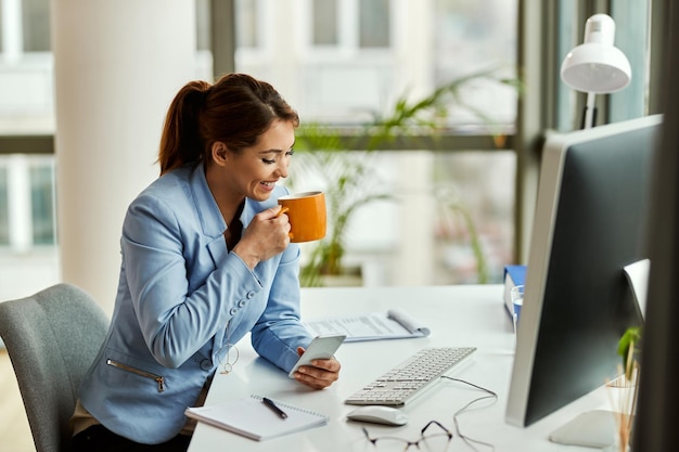 Free photo young happy businesswoman drinking coffee and text messaging on cell phone in the office