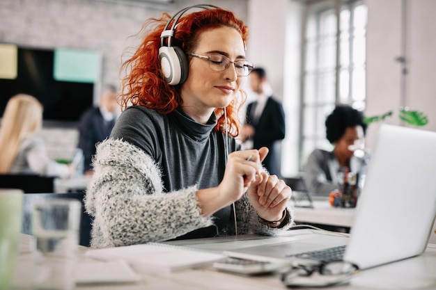 Free Photo young happy businesswoman enjoying in favorite music over headphones while working on a computer in the office there are people in the background