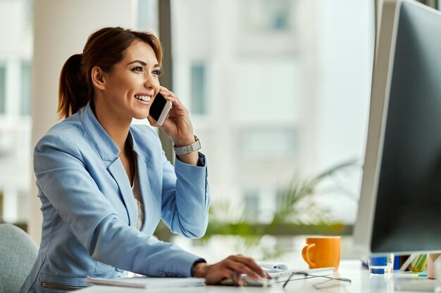 Young happy businesswoman working on desktop PC and communicating over mobile phone in the office