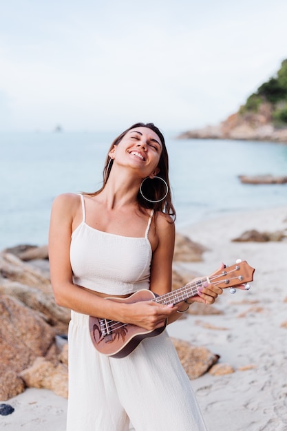 young happy calm caucasian woman with ukulele on tropical rocky beach at sunset