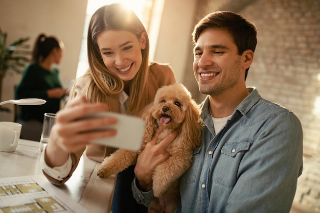 Free photo young happy colleagues having fun while taking selfie with a poodle in the office