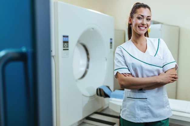 Free photo young happy doctor standing with her arms crossed next to mri scanner and looking at camera