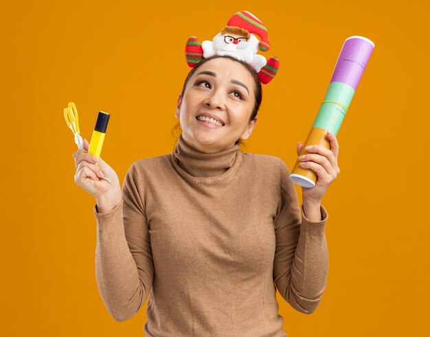 Young happy girl in funny christmas rim on head holding petard scissors and glue