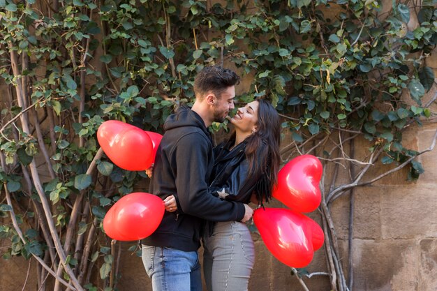 Young happy guy hugging smiling lady and holding balloons in form of hearts