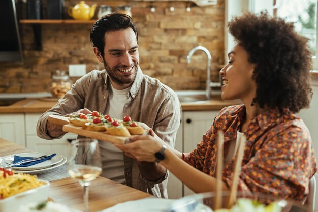 Young happy man communicating with his African American girlfriend while passing her food at dining table