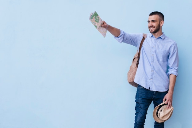 Young happy man holding map and hat showing something standing against blue background