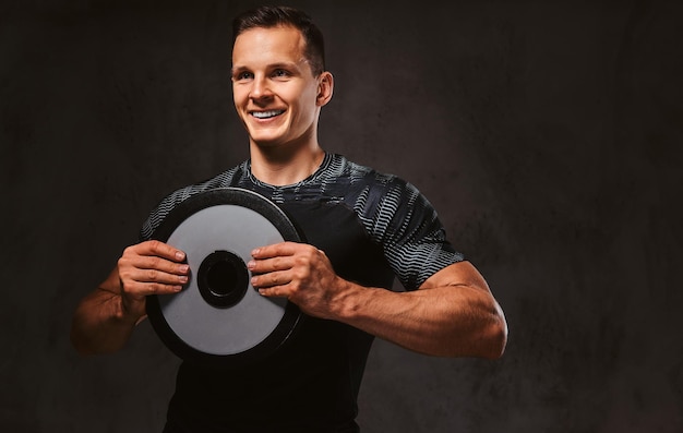 A young happy man in sportswear holding a barbell disk on a dark background.