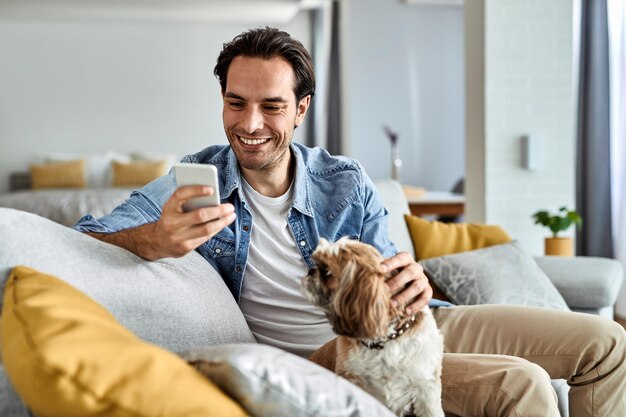 Young happy man text messaging on cell phone while sitting on the sofa with his dog