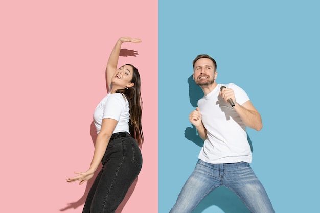 Free photo young and happy man and woman in casual clothes on pink, blue bicolored wall, singing and dancing