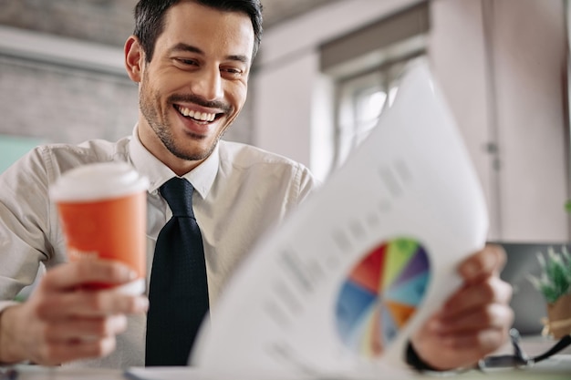 Young happy manager examining business progress while reading charts and drinking coffee in the office