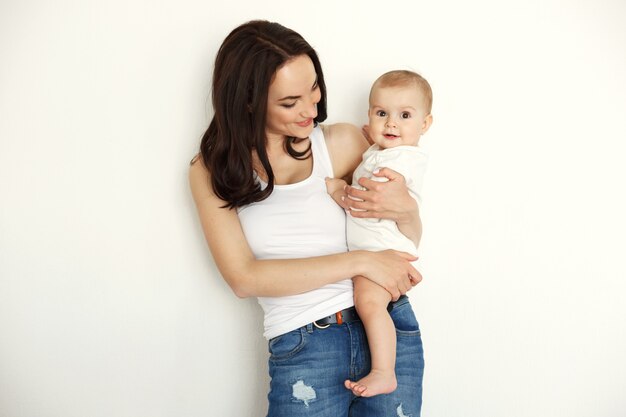 Young happy mother smiling holding looking at her baby daughter over white wall.