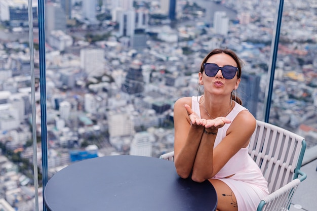 Free Photo young happy smiling caucasian woman traveller in fitting dress and sunglasses on high floor in bangkok stylish female exploring amazing big city view