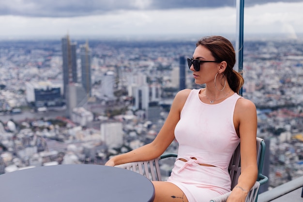 Free photo young happy smiling caucasian woman traveller in fitting dress and sunglasses on high floor in bangkok stylish female exploring amazing big city view