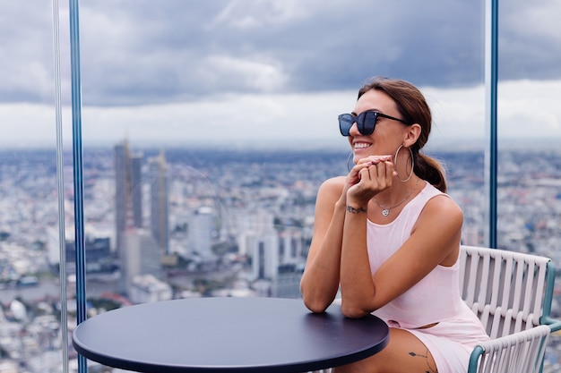 Free Photo young happy smiling caucasian woman traveller in fitting dress and sunglasses on high floor in bangkok stylish female exploring amazing big city view