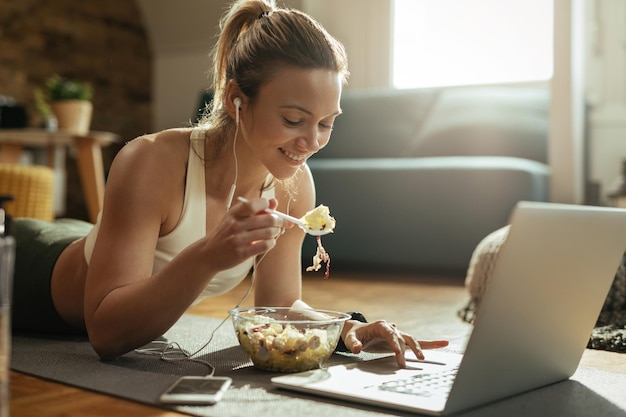 Free photo young happy sportswoman eating salad and using laptop while relaxing on the floor at home