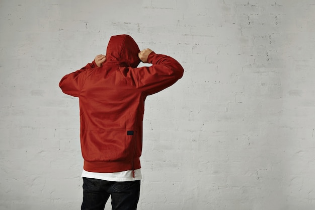 Free Photo a young hipster adjusts the hood of his brownish red parka, back view, portrait in studio with white walls