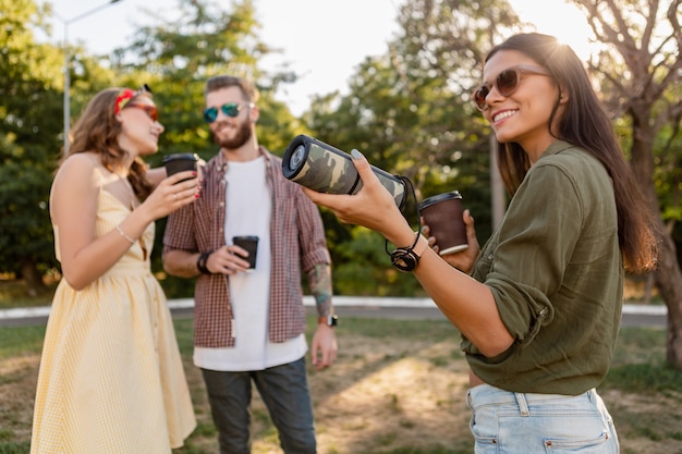 Young hipster company of friends having fun together in park smiling listening to music on wireless speaker, summer style season