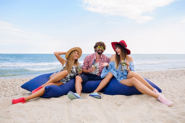 Young hipster company of friends on summer vacation sitting at beach on bean bags, having fun together, drinking mojito cocktail, happy, smiling, positive, funny emotion, three person party